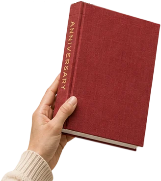 A hand holding a deep red linen hardcover photo book with ANNIVERSARY foil stamped down the spine.