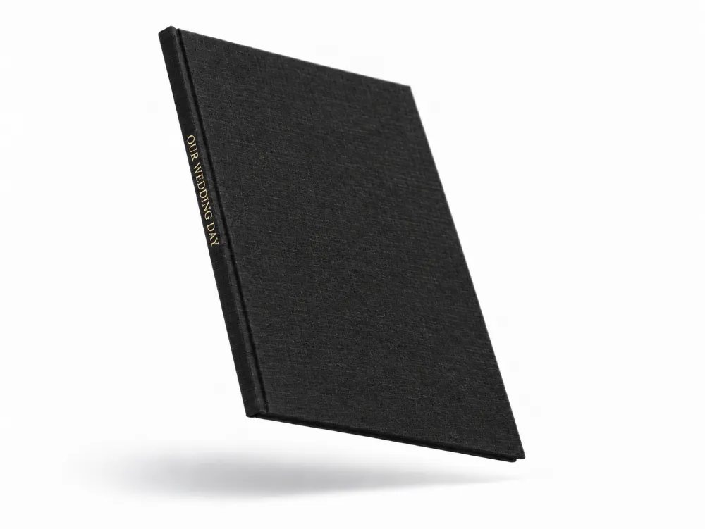 A black linen hardcover photo book floating at an angle, with OUR WEDDING DAY stamped in gold foil on the spine.