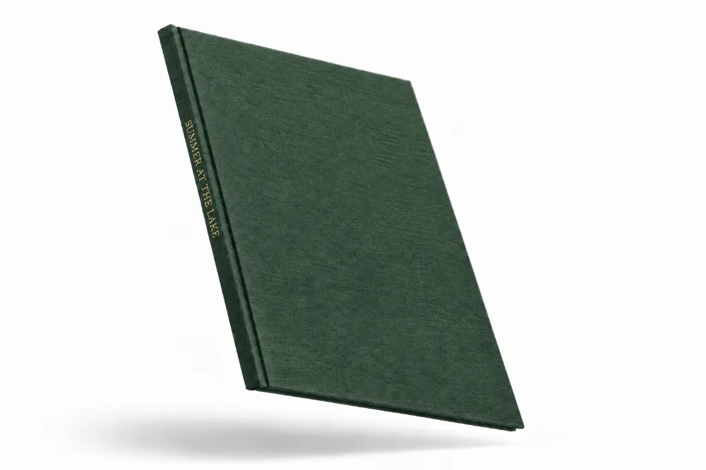 A dark green linen hardcover photo book floating at an angle, with SUMMER AT THE LAKE stamped in gold foil on the spine.