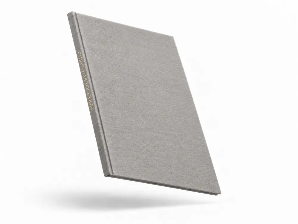 A grey linen hardcover photo book floating at an angle, with CHRISTMAS TOGETHER stamped in gold foil on the spine.