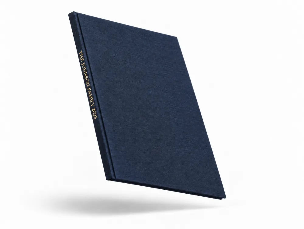 A navy linen hardcover photo book floating at an angle, with THE JOHNSON FAMILY 2025 stamped in gold foil on the spine.