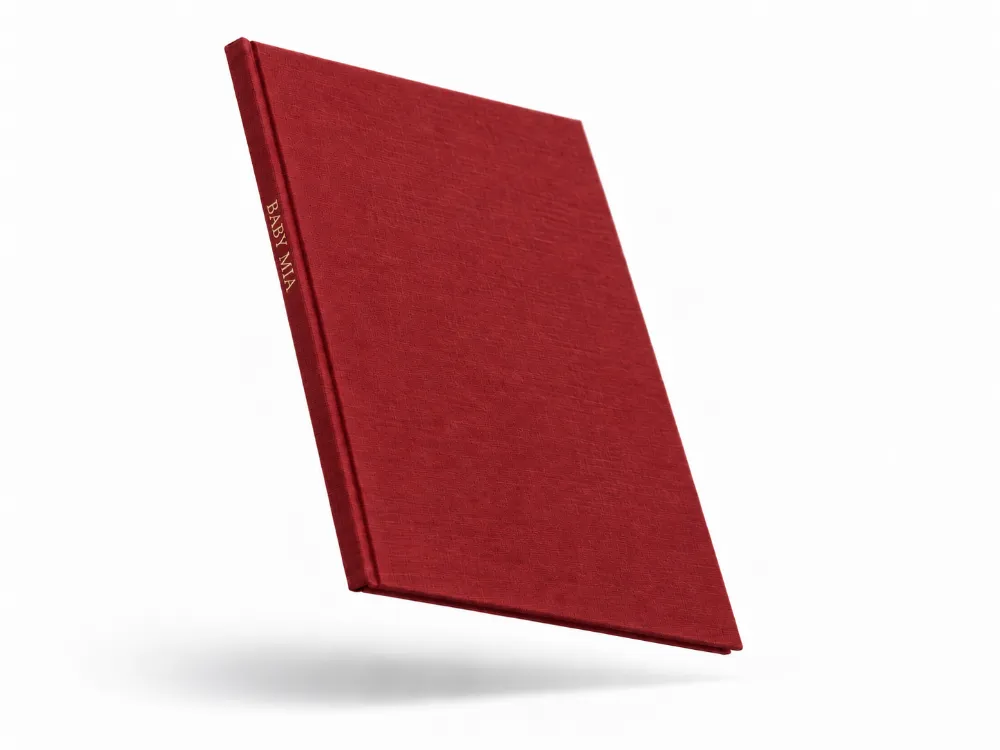 A red linen hardcover photo book floating at an angle, with BABY MIA stamped in gold foil on the spine.