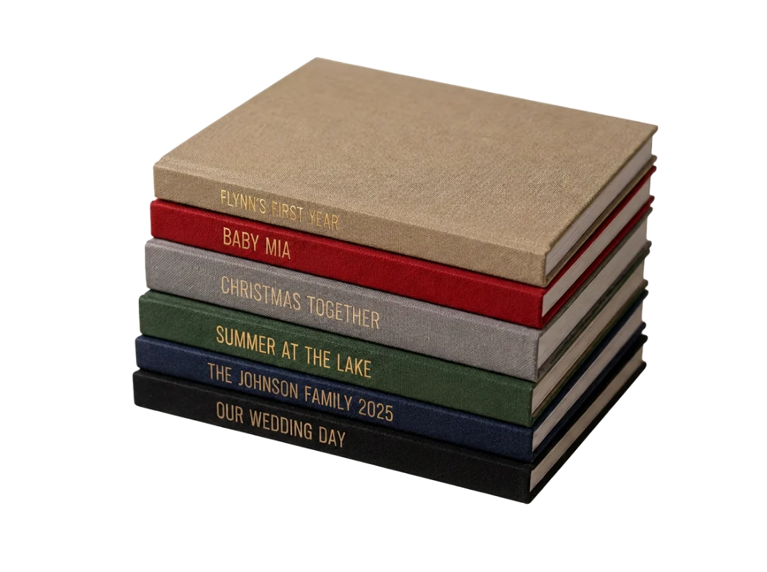 Six linen hardcover photo books stacked, in tan, red, grey, green, navy and black, each with a gold foil stamped title on the spine reading FLYNN'S FIRST YEAR, BABY MIA, CHRISTMAS TOGETHER, SUMMER AT THE LAKE, THE JOHNSON FAMILY 2025 and OUR WEDDING DAY.